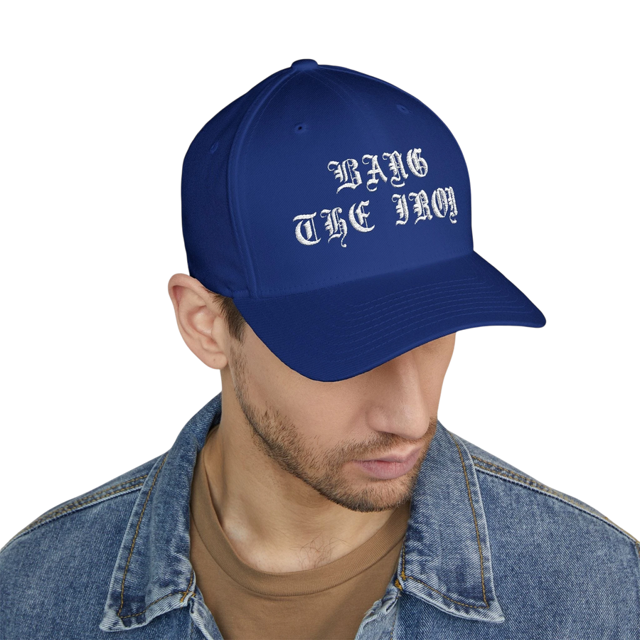 DailyThreadCo. blue structured FlexFit cap with white Bang the Iron gothic embroidery worn by a man in a denim jacket.