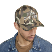 DailyThreadCo. camo FlexFit cap with white gothic Bang the Iron embroidery, worn by a man in a denim jacket.