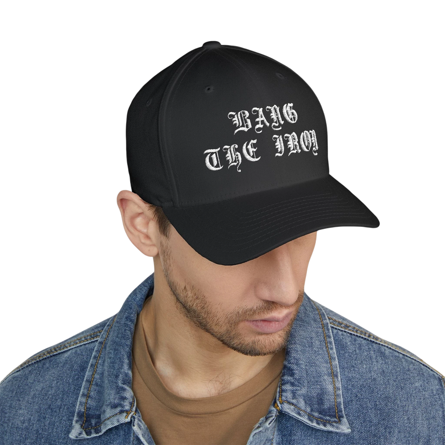 DailyThreadCo. black structured FlexFit cap with white Bang the Iron gothic embroidery worn by a man in denim jacket.