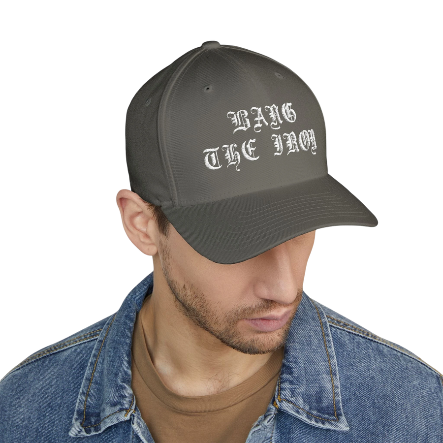 DailyThreadCo. structured grey FlexFit cap with white Bang the Iron gothic embroidery worn by a man in a denim jacket.