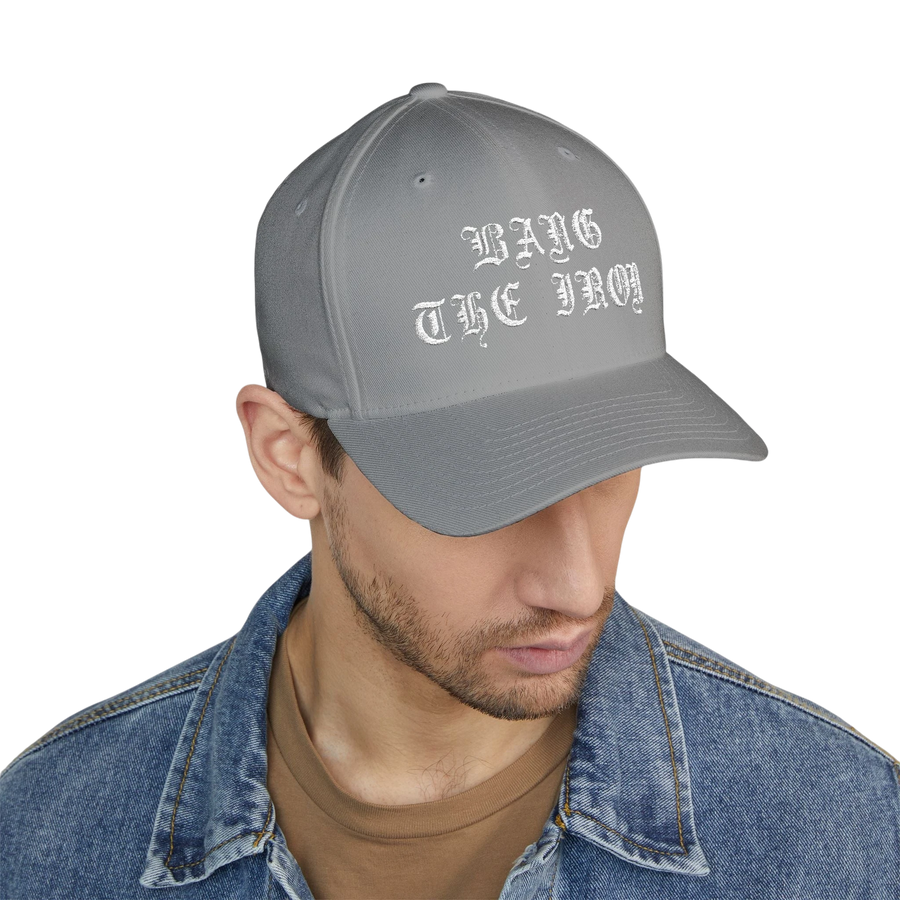 DailyThreadCo. grey structured FlexFit cap with white gothic Bang the Iron embroidery worn by a man in a denim jacket.